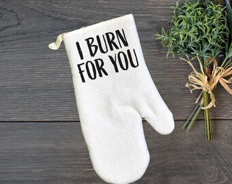 May include: White oven mitt with the text "I BURN FOR YOU" in black. A bundle of green and purple faux plants tied with twine sits to the right. The items are on a dark wood surface.