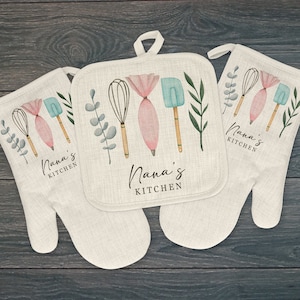 May include: A set of two white cotton oven mitts and a pot holder with a floral design. The mitts and pot holder feature a watercolor illustration of kitchen utensils, including a whisk, spatula, rolling pin, and a sprig of eucalyptus. The text "Nana's Kitchen" is printed on each item.