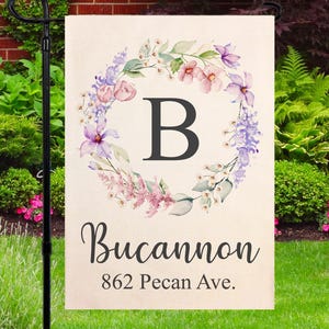 May include: A garden flag with a floral wreath design. The flag features a large black letter "B" in the center, surrounded by a wreath of pink, purple, and white flowers. The name "Bucannon" and address "862 Pecan Ave." are below.