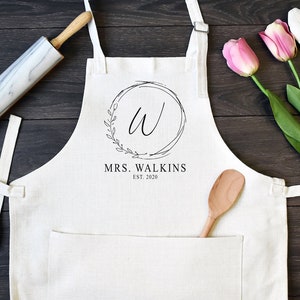 May include: White apron with a large pocket, featuring a black and white design of a wreath with the letter "W" inside and the text "MRS. WALKINS EST. 2020".