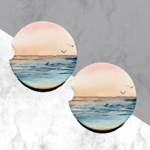 May include: Two round ceramic coasters with a watercolor design of a sunset over the ocean. The coasters feature a light blue ocean with a light orange sunset in the background. There are small black birds flying in the sky.