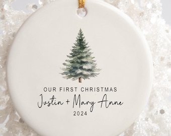 May include: A white ceramic Christmas ornament with a watercolor pine tree design. The text "OUR FIRST CHRISTMAS" is above the names "Justin + Mary Anne" and the year "2024" in a script font. The ornament hangs from a gold cord.