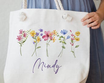 May include: White canvas tote bag with a floral design and the name "Mindy" printed in purple. The flowers are in shades of pink, yellow, blue, and purple.