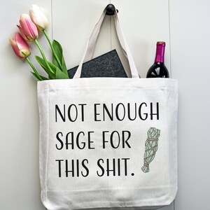 May include: White canvas tote bag with black text that reads "Not Enough Sage For This Shit." A small illustration of a bundle of sage is to the right of the text. A bottle of wine and a bouquet of pink tulips are in the bag.