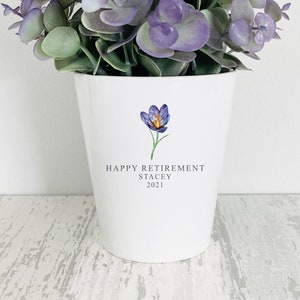 May include: White ceramic planter with a purple flower graphic and the text "Happy Retirement Stacey 2021". The planter is filled with a small artificial eucalyptus plant with purple leaves.