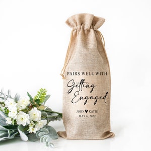 Op de afbeelding: Een jute wijnfleszak met de tekst "Pairs well with Getting Engaged John &hearts; Katie May 6, 2022".
