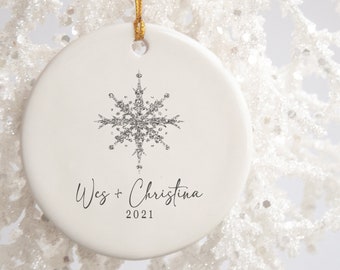 May include: White ceramic Christmas ornament with a silver glitter snowflake and the text "Wes + Christina 2021".