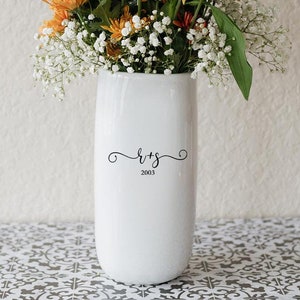 May include: A white ceramic vase with a floral arrangement of orange, yellow, and white flowers. The vase has a curved design and features the initials "R+S" and the year "2003" in black script.