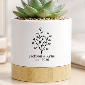 May include: A white cylindrical ceramic planter with a light wood base containing a green succulent. The front of the planter features a black line drawing of a leafy branch above the printed text Jackson + Kylie est. 2026 in black font.