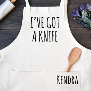 May include: A cream-colored kitchen apron featuring the black text I'VE GOT A KNIFE printed on the chest and the name KENDRA printed on the front pocket. The apron includes a neck strap and two waist ties, with a marble rolling pin and wooden spoon.