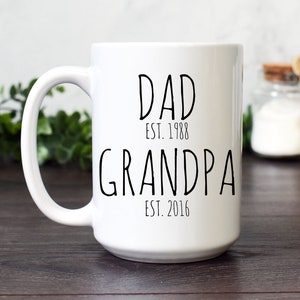Może przedstawiać: Biały ceramiczny kubek z czarnym napisem "DAD EST. 1988 GRANDPA EST. 2016". Kubek ma duży uchwyt i stoi na ciemnej drewnianej powierzchni. W tle dekoracyjne słoiki i zieleń.