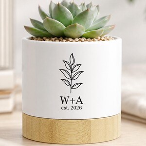 May include: A white cylindrical ceramic planter with a light wood base, containing a green succulent. The front features a black line drawing of a leafy branch above the text W+A and est. 2026 in a clean, black serif font centered on the pot.