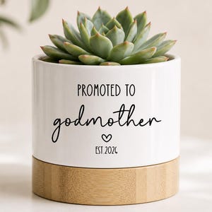 May include: A white ceramic cylindrical planter with a light wood base containing a green succulent. The planter features black text that reads PROMOTED TO godmother with a small heart icon and the text EST. 2026 printed on the front surface.
