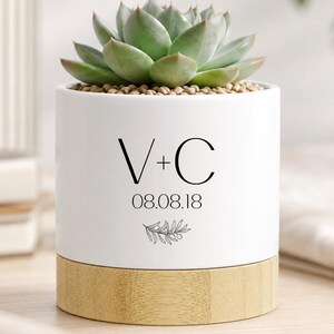 May include: A white ceramic planter with a bamboo base, holding a green succulent. The planter is decorated with the initials "V+C", the date "08.08.18", and a small leaf design. A decorative item for the home.