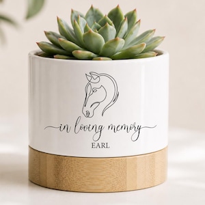 May include: A white ceramic planter with a line drawing of a horse's head and the text "in loving memory EARL". The planter has a natural wood base and is filled with a green succulent plant.