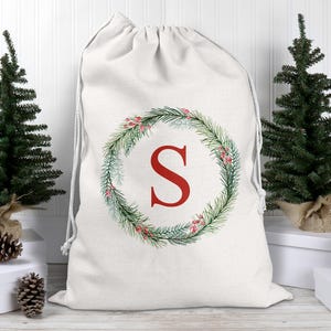 May include: A white drawstring Christmas gift bag with a red letter "S" inside a green wreath with red berries. Two small Christmas trees and pinecones flank the bag, set against a white background.