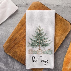 May include: White waffle-weave kitchen towel featuring a watercolor Christmas tree design with presents at the base. The text "The Treys" is written in a script font. The towel is on a wooden cutting board.