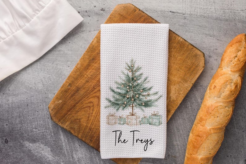 May include: White waffle-weave kitchen towel featuring a watercolor Christmas tree design with presents at the base. The text "The Treys" is written in a script font. The towel is on a wooden cutting board.