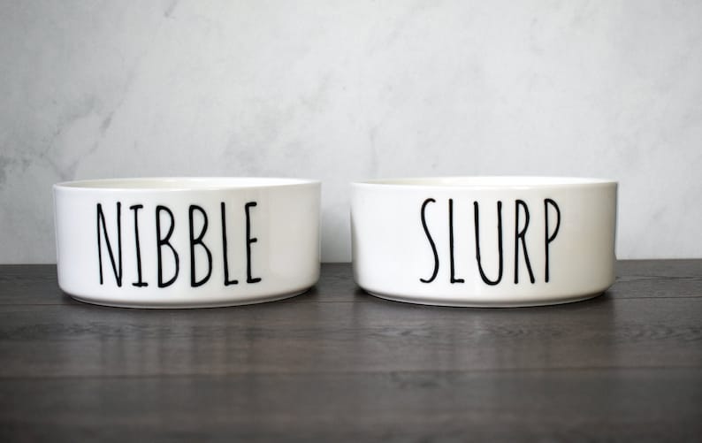 May include: Two white ceramic pet bowls with black lettering. The bowl on the left says "Nibble" and the bowl on the right says "Slurp".