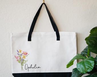 May include: A white canvas tote bag with black handles and a black bottom. The bag has a floral design and the name "Ophelia" printed on the front.