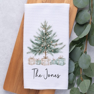 May include: White waffle-weave kitchen towel featuring a watercolor Christmas tree design with gold ornaments and wrapped gifts. The text "The Jones" is written in a script font below the gifts. The towel is on a wooden cutting board.