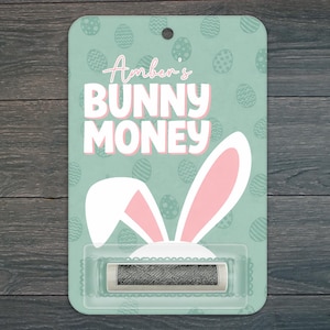 May include: A rectangular light green card with a repeating egg pattern, featuring the text Amber's BUNNY MONEY in white and pink. A white and pink bunny ear graphic sits above a clear plastic blister pack containing a rolled silver coin.