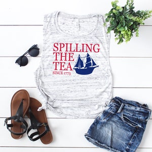 K&ouml;nnte beinhalten: Ein &auml;rmelloses Tanktop mit Marmormuster und dem Text "Spilling the Tea Since 1773" in Rot und Marineblau. Das Bild zeigt auch eine schwarze Sonnenbrille, braune Sandalen und Jeansshorts, angeordnet auf einer wei&szlig;en Holzoberfl&auml;che.