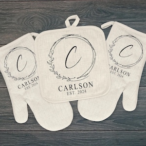May include: Set of three beige kitchen pot holders with black embroidery. The pot holders feature a wreath design with a letter C in the center and the text "CARLSON EST. 2024".