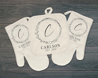 May include: Set of three beige kitchen pot holders with black embroidery. The pot holders feature a wreath design with a letter C in the center and the text "CARLSON EST. 2024".