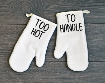 May include: A pair of white oven mitts with black text. One mitt says "Too Hot" and the other says "To Handle".