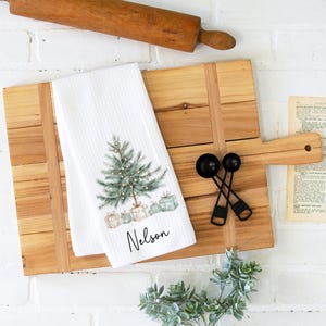May include: A white kitchen towel with a Christmas tree design and the name "Nelson" printed on it. The towel is on a wooden cutting board with a rolling pin, measuring spoons, and a vintage recipe page.