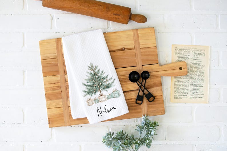 May include: A white kitchen towel with a Christmas tree design and the name "Nelson" printed on it. The towel is on a wooden cutting board with a rolling pin, measuring spoons, and a vintage recipe page.