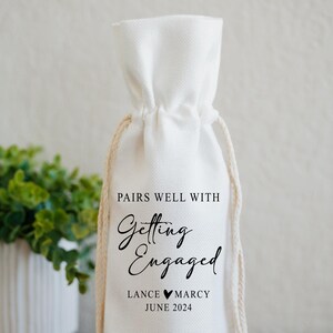 Op de afbeelding: Een witte katoenen wijnzak met de tekst "Pairs well with Getting Engaged Lance &hearts; Marcy June 2024"