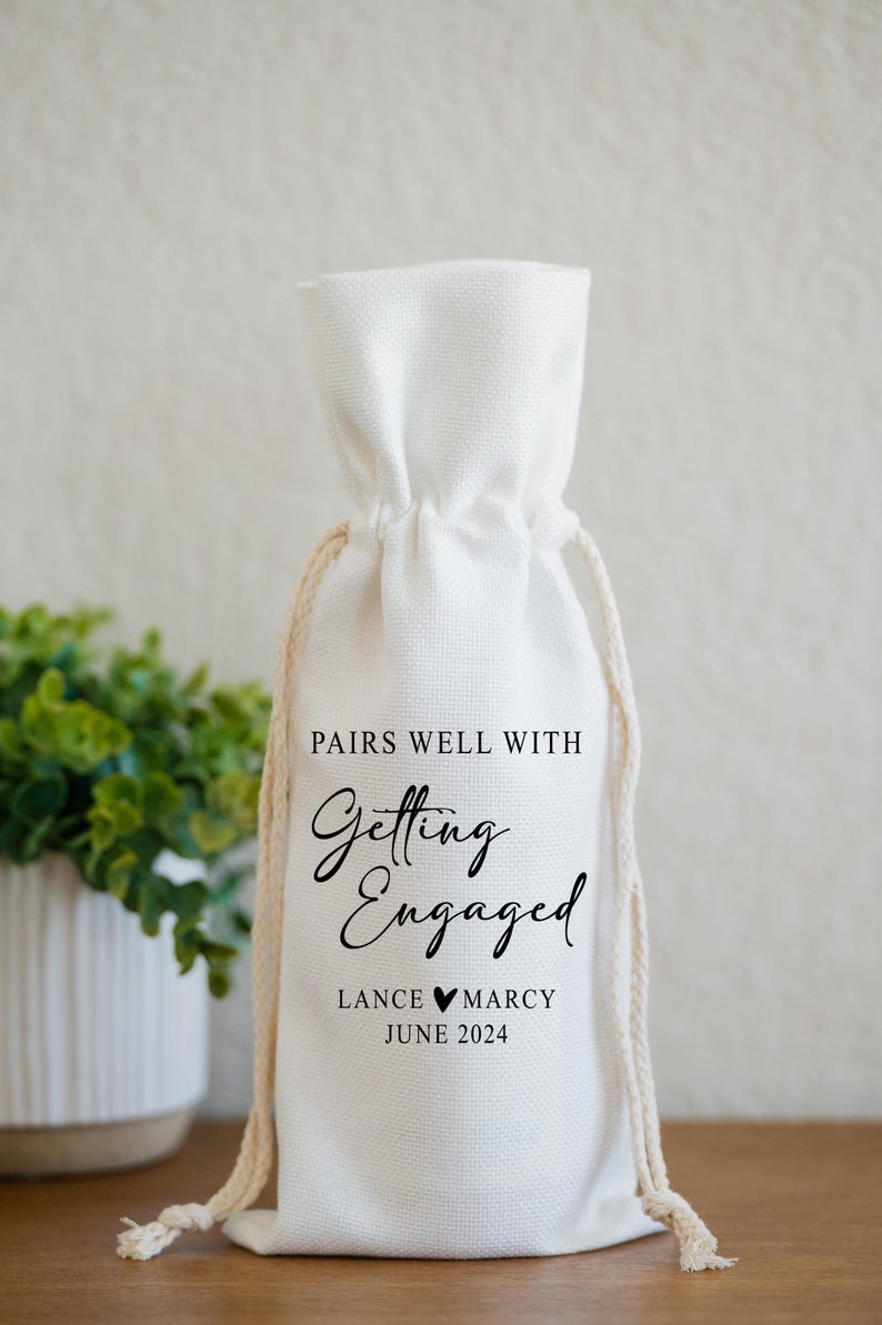 Op de afbeelding: Een witte katoenen wijnzak met de tekst "Pairs well with Getting Engaged Lance &hearts; Marcy June 2024"