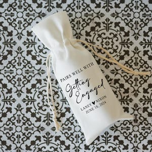 Op de afbeelding: Een witte stoffen cadeauzak met een zwart bedrukt etiket met de tekst "Pairs well with Getting Engaged", de namen "Laney & Nixon" en de datum "June 18, 2024".