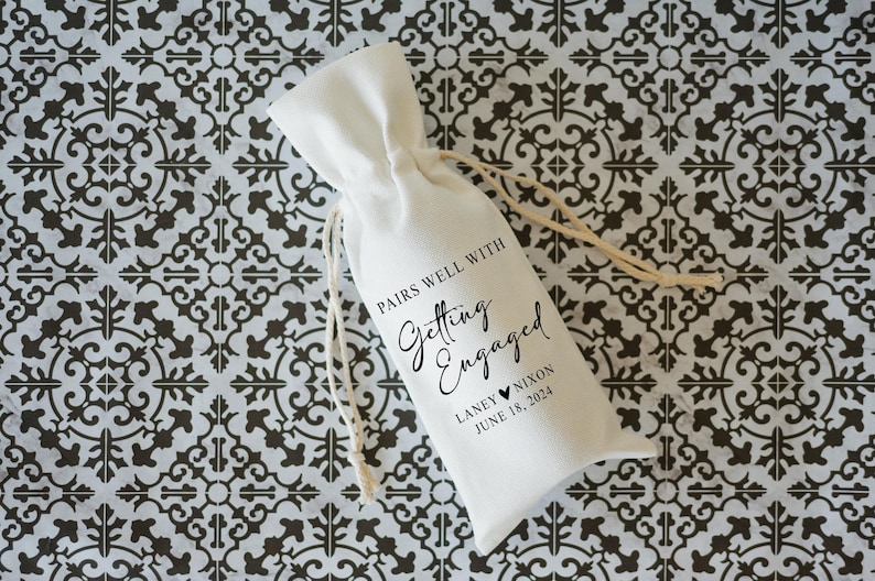 Op de afbeelding: Een witte stoffen cadeauzak met een zwart bedrukt etiket met de tekst "Pairs well with Getting Engaged", de namen "Laney & Nixon" en de datum "June 18, 2024".