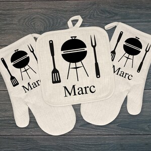 May include: A set of three personalized barbecue-themed kitchen textiles. The set includes two oven mitts and a square pot holder. Each item is off-white with a black graphic of a grill and the name "Marc".