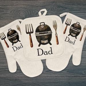 May include: A set of three off-white kitchen textiles featuring a watercolor design. The set includes two oven mitts and a square pot holder. Each item displays a black grill, a fork, and the word "Dad" in black text.