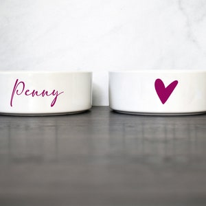 Puede incluir: Dos cuencos de cer&aacute;mica blanca para mascotas. Un cuenco tiene el nombre "Penny" escrito en cursiva rosa. El otro cuenco tiene un coraz&oacute;n rosa.