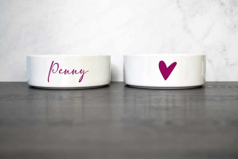 Puede incluir: Dos cuencos de cer&aacute;mica blanca para mascotas. Un cuenco tiene el nombre "Penny" escrito en cursiva rosa. El otro cuenco tiene un coraz&oacute;n rosa.
