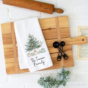 May include: White kitchen towel with a watercolor Christmas tree design, gifts, and the text "The Traxon Family." The towel is on a wooden cutting board with a rolling pin, measuring spoons, and a vintage book.