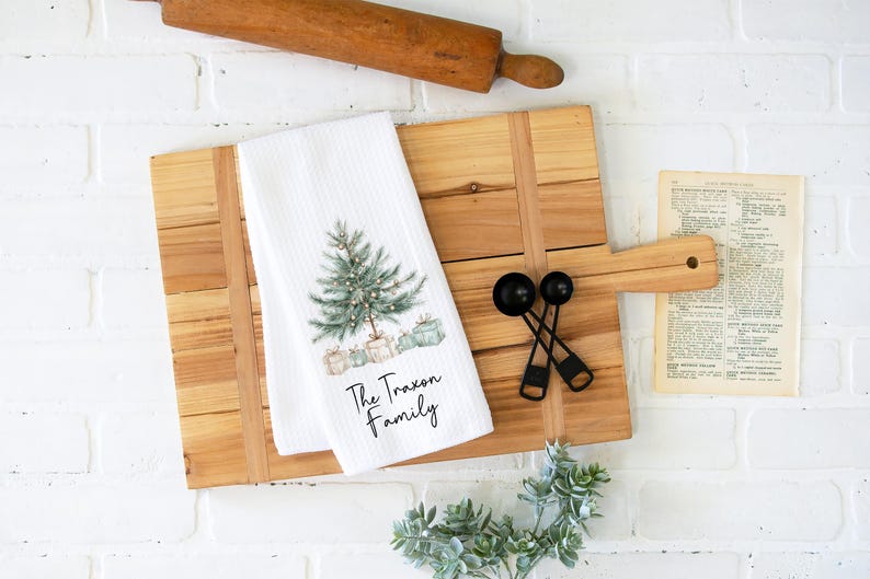 May include: White kitchen towel with a watercolor Christmas tree design, gifts, and the text "The Traxon Family." The towel is on a wooden cutting board with a rolling pin, measuring spoons, and a vintage book.