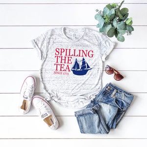 K&ouml;nnte beinhalten: Ein meliertes graues T-Shirt mit dem Text "Spilling the Tea Since 1773" und einer blauen Segelboot-Grafik. Das Shirt wird mit einer blauen Jeans, wei&szlig;en Turnschuhen und einer Sonnenbrille kombiniert. Ein Zweig Gr&uuml;nzeug ist ebenfalls vorhanden.