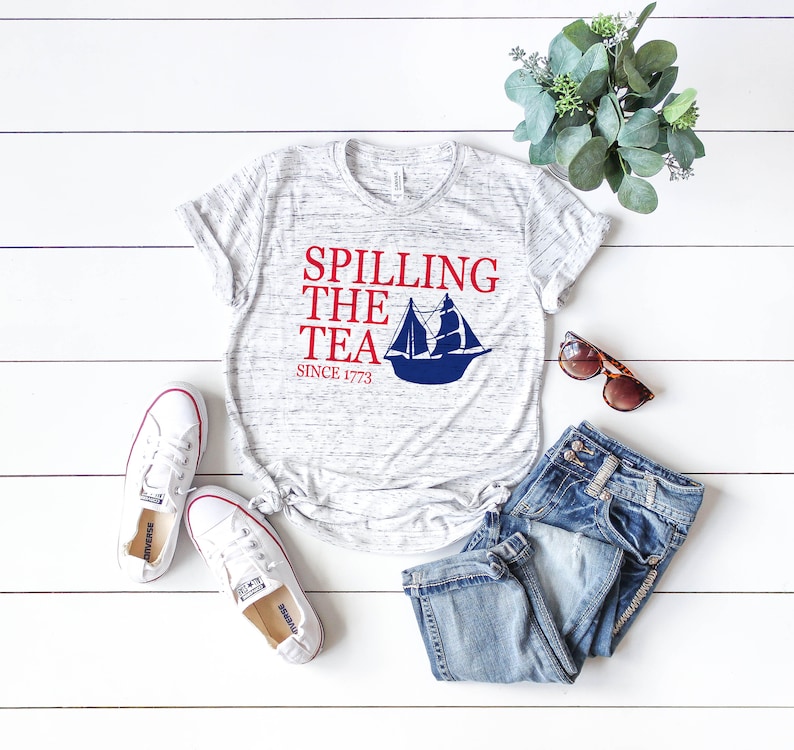 K&ouml;nnte beinhalten: Ein meliertes graues T-Shirt mit dem Text "Spilling the Tea Since 1773" und einer blauen Segelboot-Grafik. Das Shirt wird mit einer blauen Jeans, wei&szlig;en Turnschuhen und einer Sonnenbrille kombiniert. Ein Zweig Gr&uuml;nzeug ist ebenfalls vorhanden.