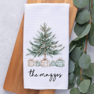 May include: White waffle-weave kitchen towel featuring a watercolor Christmas tree design with presents and the text "the mazzos" in black script. The towel is displayed on a wooden cutting board.