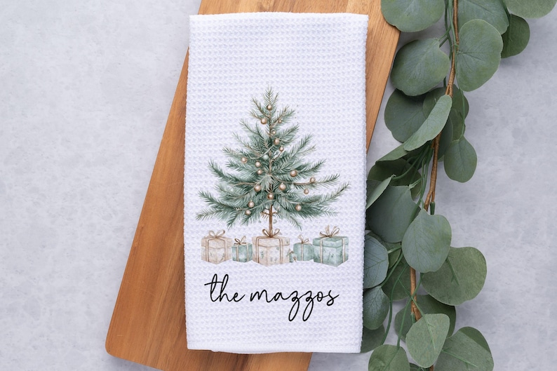May include: White waffle-weave kitchen towel featuring a watercolor Christmas tree design with presents and the text "the mazzos" in black script. The towel is displayed on a wooden cutting board.