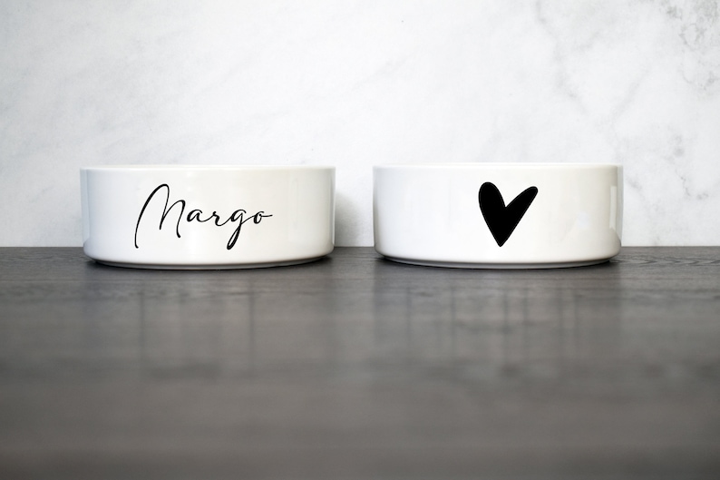 Puede incluir: Dos cuencos de cer&aacute;mica blanca para mascotas, uno con el nombre "Margo" impreso en letra negra y el otro con un dise&ntilde;o de coraz&oacute;n negro.