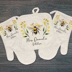 May include: A set of kitchen linens featuring a watercolor design of a bumblebee and yellow flowers. The set includes two oven mitts and a square pot holder. The text "Mrs. Daniel's Kitchen" is printed on each item.