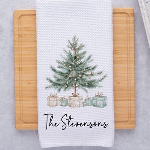 May include: White waffle-weave kitchen towel featuring a watercolor Christmas tree design with gifts. The text "The Stevensons" is printed in a script font below the tree. The towel is on a wooden cutting board.