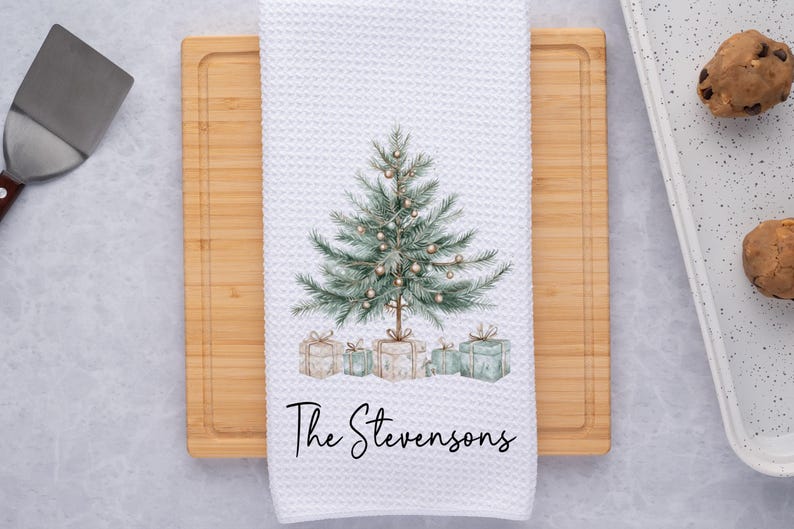 May include: White waffle-weave kitchen towel featuring a watercolor Christmas tree design with gifts. The text "The Stevensons" is printed in a script font below the tree. The towel is on a wooden cutting board.